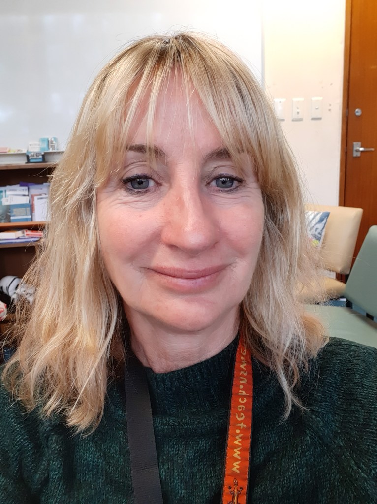 Libby Purcell. Registered Psychologist, Vida Psychology Support. Close-up selfie of a woman with shoulder-length blonde hair, wearing a green sweater and a colorful lanyard, with a classroom background.